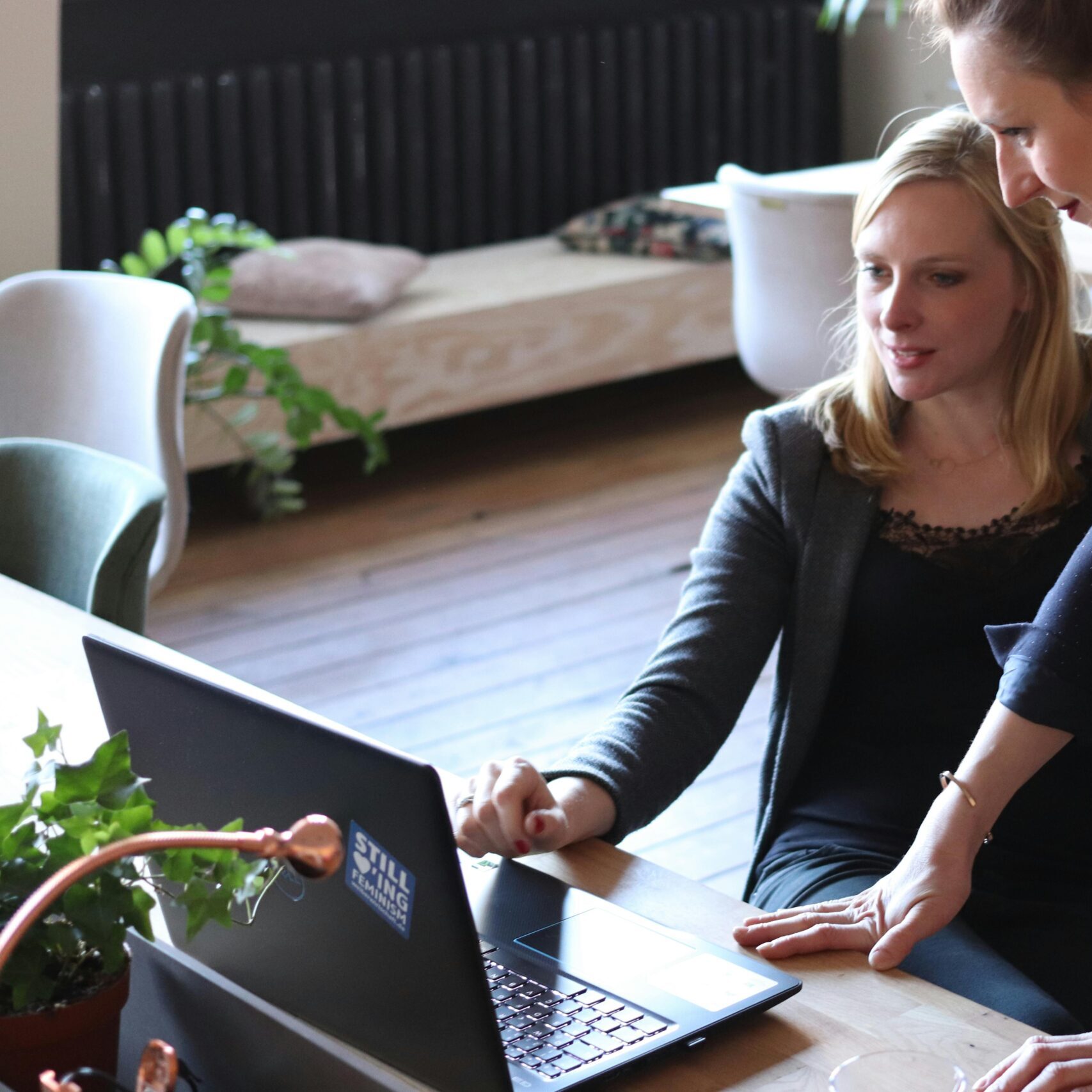 Two professional women discuss a project using a laptop in a modern office environment.